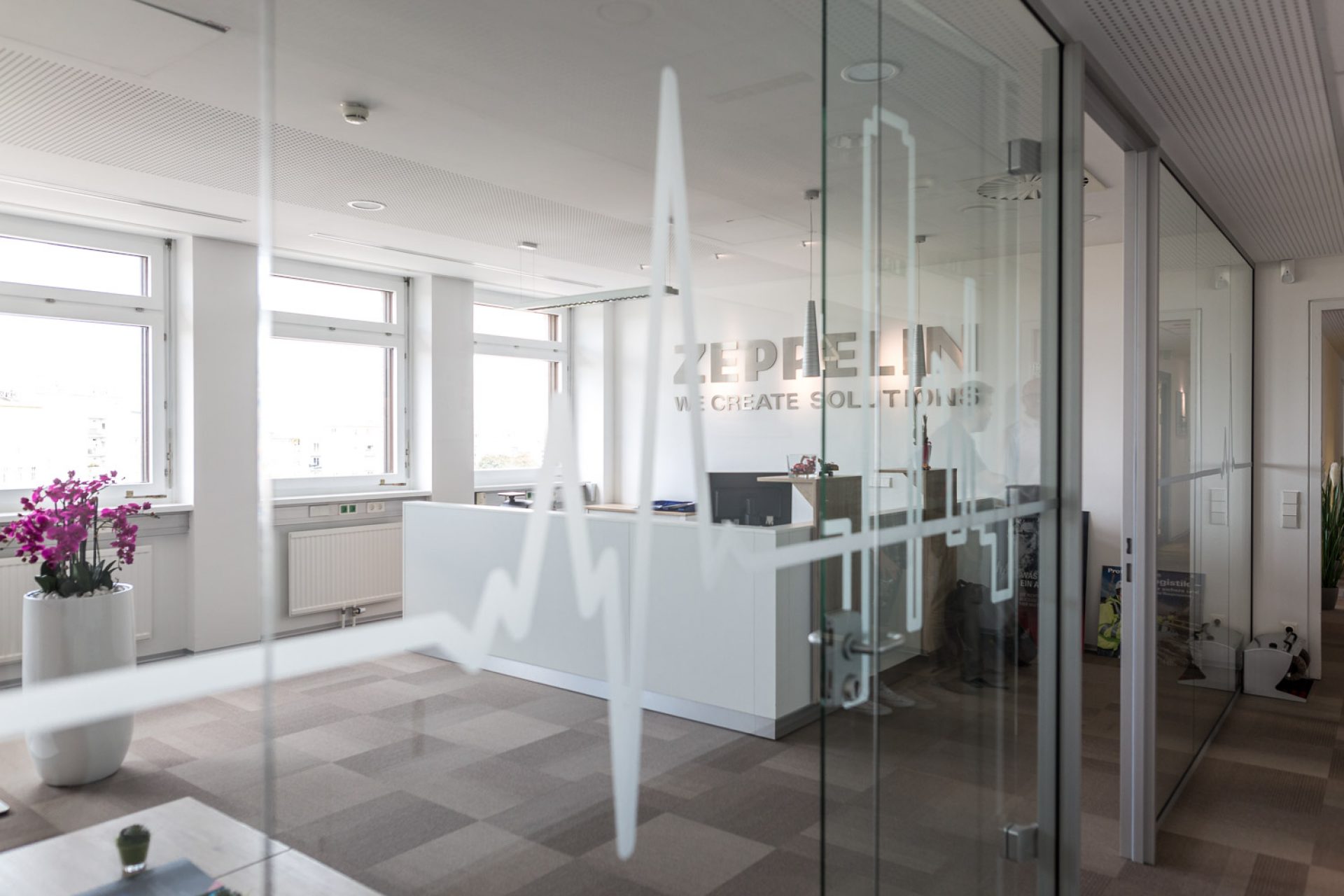 Büro mit Glastür, „ZEPPELIN WE CREATE SOLUTIONS“ darauf, Schreibtisch und PC im Hintergrund, Orchidee im Blumentopf links.