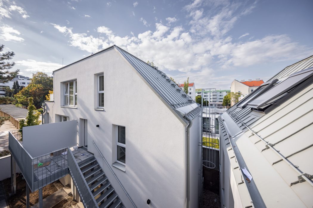 Zweistöckiges modernes Gebäude mit weißer Fassade, Metalldach und Außentreppe, umgeben von Bäumen und Nachbarhäusern im Hintergrund.