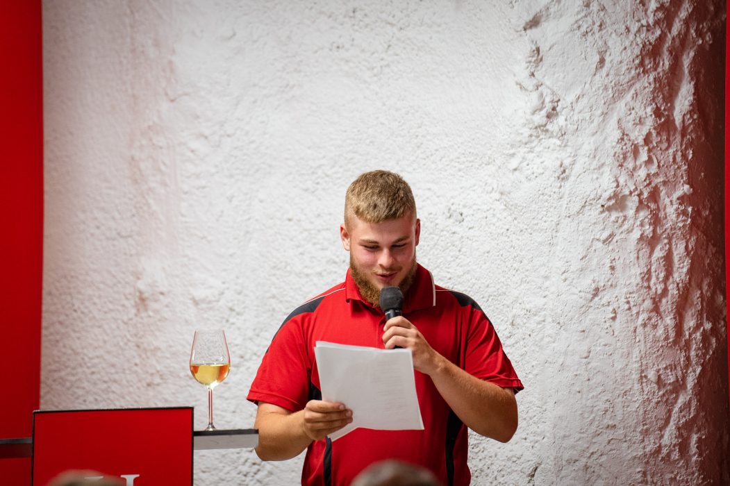 Ein Mann in einem roten Hemd hält ein Mikrofon und liest von einem Blatt Papier, während ein Glas mit Getränk auf einem Tisch neben ihm steht.