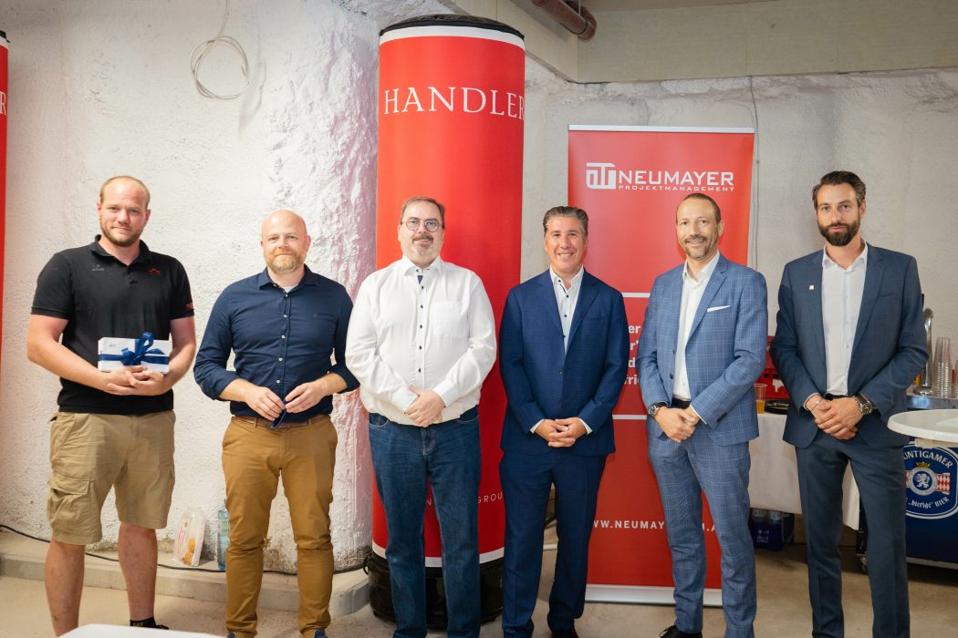 Sechs Männer in Business-Kleidung stehen in einer Reihe vor roten Bannern mit den Logos von "HANDLER" und "NEUMAYER PROJEKTMANAGEMENT".