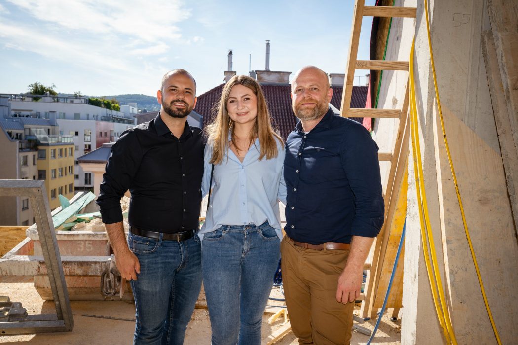 Drei Personen stehen auf einer Baustelle, eine Frau in der Mitte, umgeben von zwei Männern. Im Hintergrund sind Gebäude und ein Dach zu sehen.