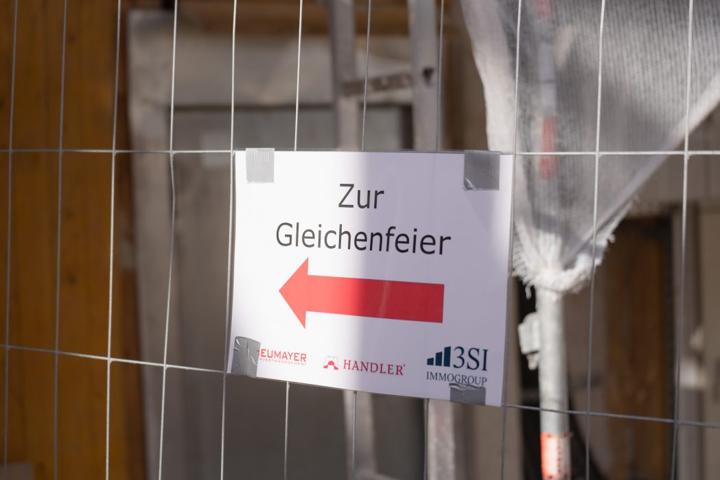 Ein Schild mit Pfeil nach links und dem Text "Zur Gleichenfeier" ist an einem Bauzaun befestigt. Logos von Firmen sind unten sichtbar.