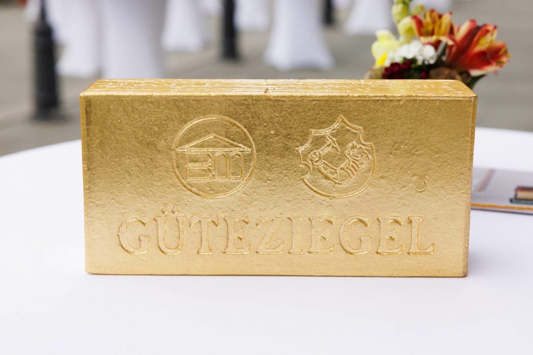 Ein goldener Ziegelstein mit den Prägungen eines Hauses, eines Wappens und dem Wort "GÜTEZIEGEL" steht auf einem weißen Tisch.