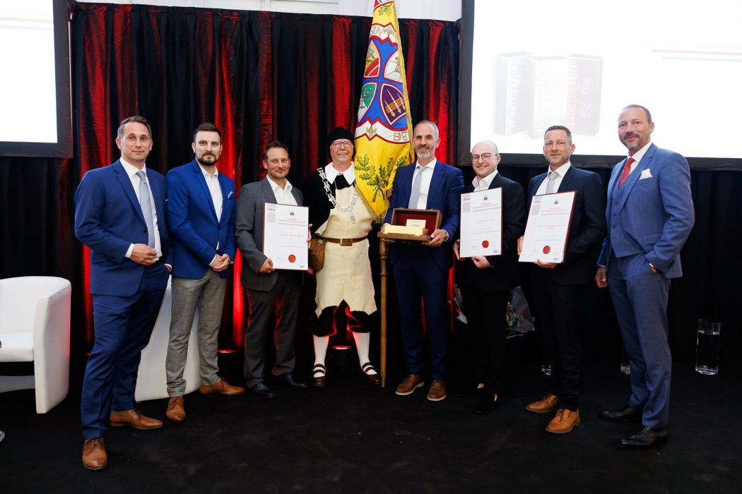 Gruppe von acht Männern in Anzügen posiert mit Urkunden und einer Trophäe vor einem Bühnenhintergrund, einer Mann in traditionellem Anzug.