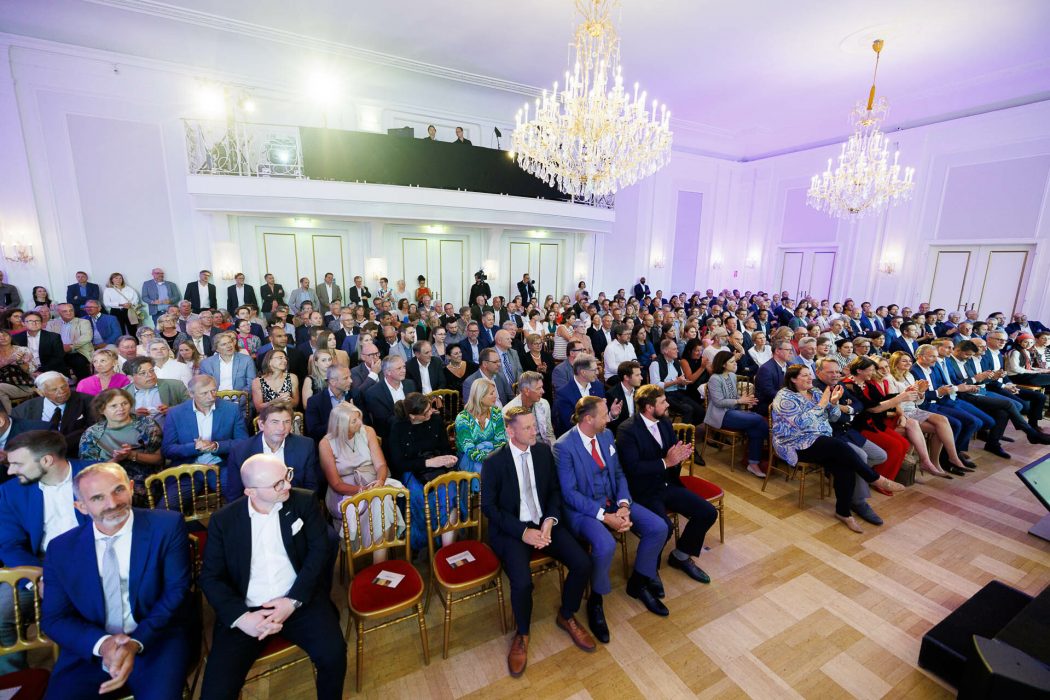 Menschenmenge in elegantem Saal mit Kronleuchtern, viele sitzen und klatschen, einige stehen hinten; formelle Kleidung, festliche Atmosphäre.