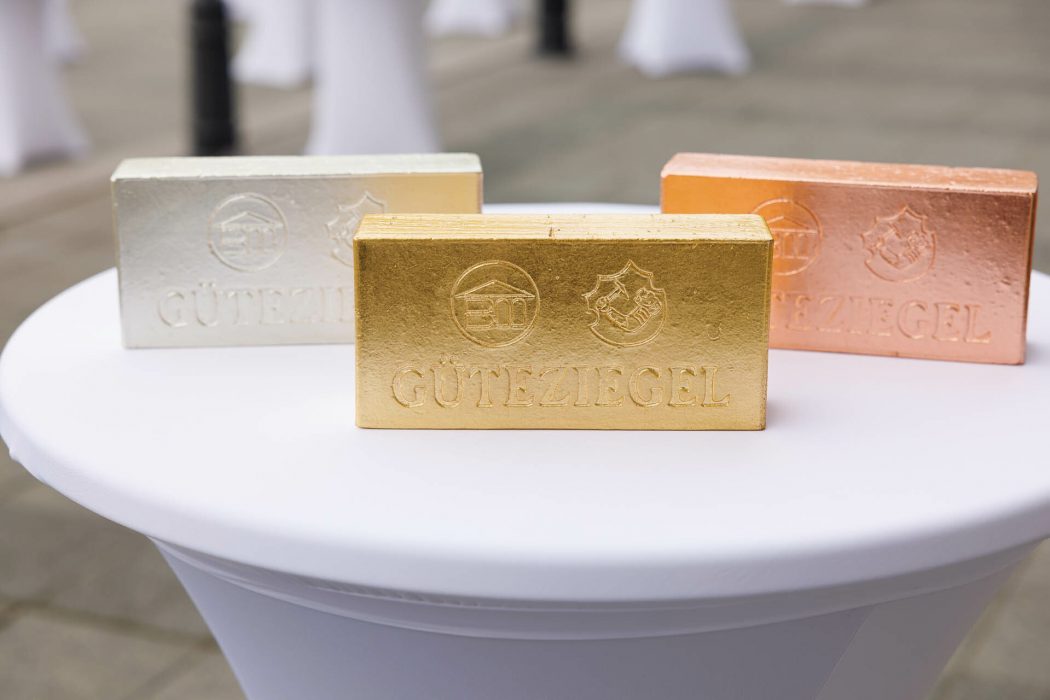 Drei dekorative Mauersteine in Gold, Silber und Bronze mit Prägung "GÜTEZIEGEL" auf einem runden weißen Tisch.