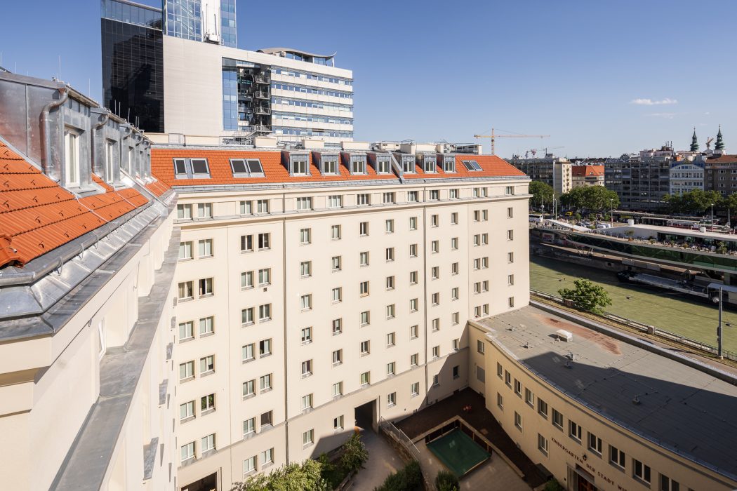 Mehrstöckiges Gebäude mit rotem Dach vor einem Fluss, im Hintergrund moderne und historische Stadtarchitektur sowie blauer Himmel.