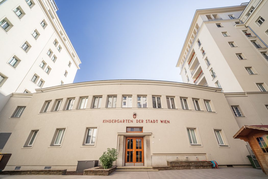 Fassade eines Kindergartens mit der Aufschrift "Kindergarten der Stadt Wien", flankiert von zwei hohen Wohngebäuden unter klarem Himmel.