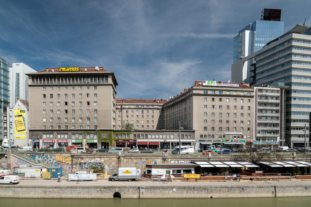 Großes Gebäude mit dem Schriftzug "Kosmos", Fluss im Vordergrund, Graffiti an der Uferwand, Fahrzeuge und Menschen auf der Straße.