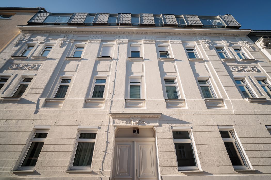 Fassade eines vierstöckigen weißen Gebäudes mit mehreren Fenstern, dekorativen Details und einem Dach mit Schindeln und Glasbalkonen.