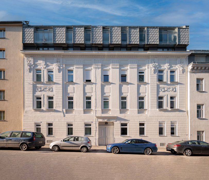 Ein weißes, mehrstöckiges Gebäude mit klassischer Fassade und modernen Dachgauben; Autos parken davor auf der Straße.