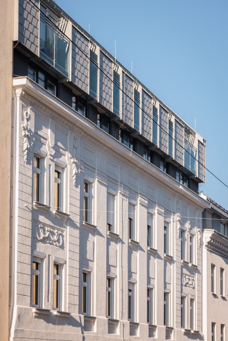 Weiße Gebäudefassade im klassischen Stil, darüber moderne blau-graue Dachaufbauten mit großen Fenstern und Schindeln. Blauer Himmel.
