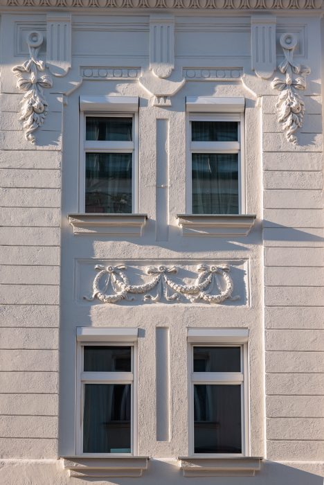 Fassade eines weißen Gebäudes mit vier Fenstern und dekorativen Verzierungen, darunter florale Muster und Reliefs in symmetrischem Design.