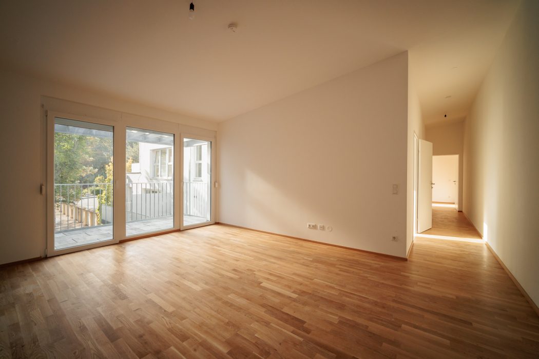 Leerer Raum mit Holzboden, großen Glas-Schiebetüren, die zu einem Balkon führen, und einem offenen Durchgang zu einem hellen Flur.