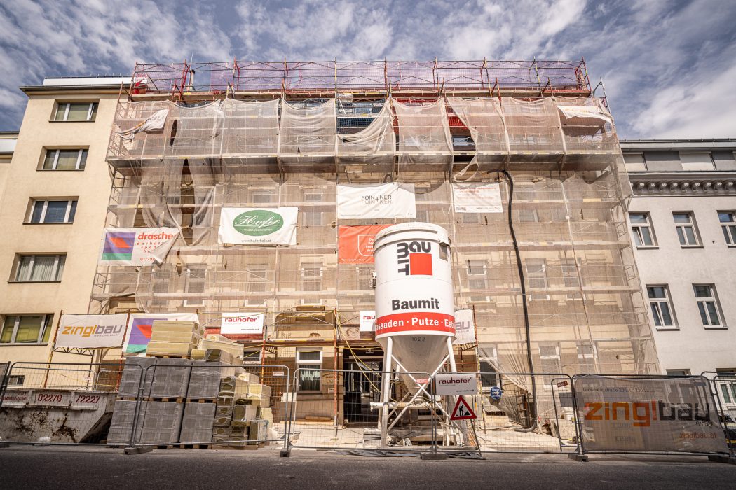 Ein Gebäude wird renoviert, eingerüstet und mit Netzen abgedeckt, vorne sind Baumaterialien und ein großer Baumit-Mischer zu sehen.