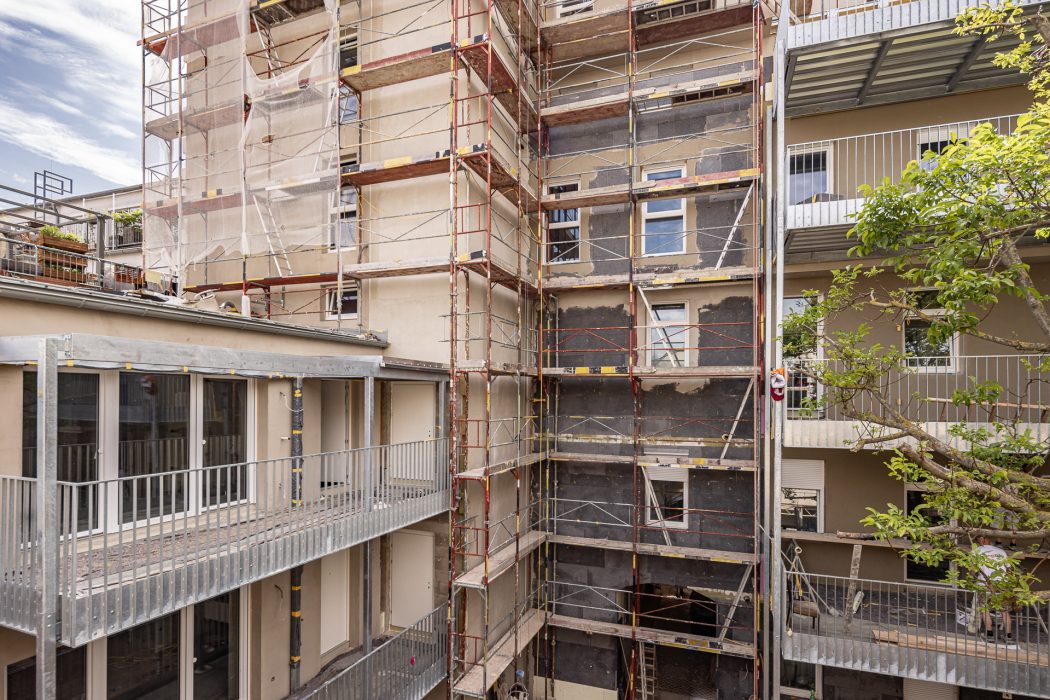 Mehrstöckiges Gebäude im Bau mit Gerüsten und Netzen, sichtbare Türen und Fensterfronten, Bäume im Vordergrund.
