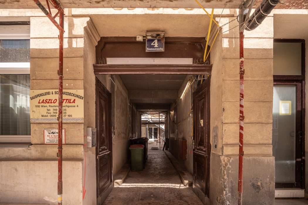 Ein schmaler Durchgang in einem Gebäude mit der Adresse "Rautenstrauchgasse 4", links Schilder mit Kontaktdaten und rechts eine offene Tür.