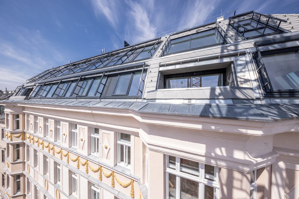Das Bild zeigt die Fassade eines mehrstöckigen modernen Gebäudes mit großen Dachfenstern und verzierten Elementen, vor einem blauen Himmel.