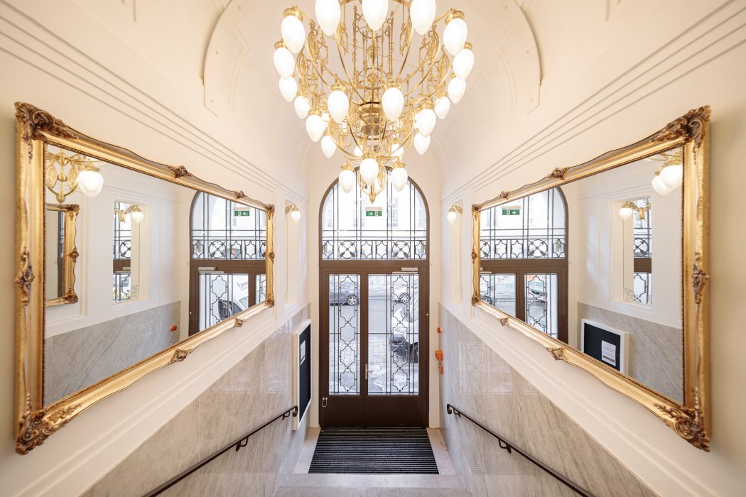 Treppenhaus mit goldenen Spiegelrahmen und einem großen, eleganten Kronleuchter, der über dem zentralen Eingang hängt.