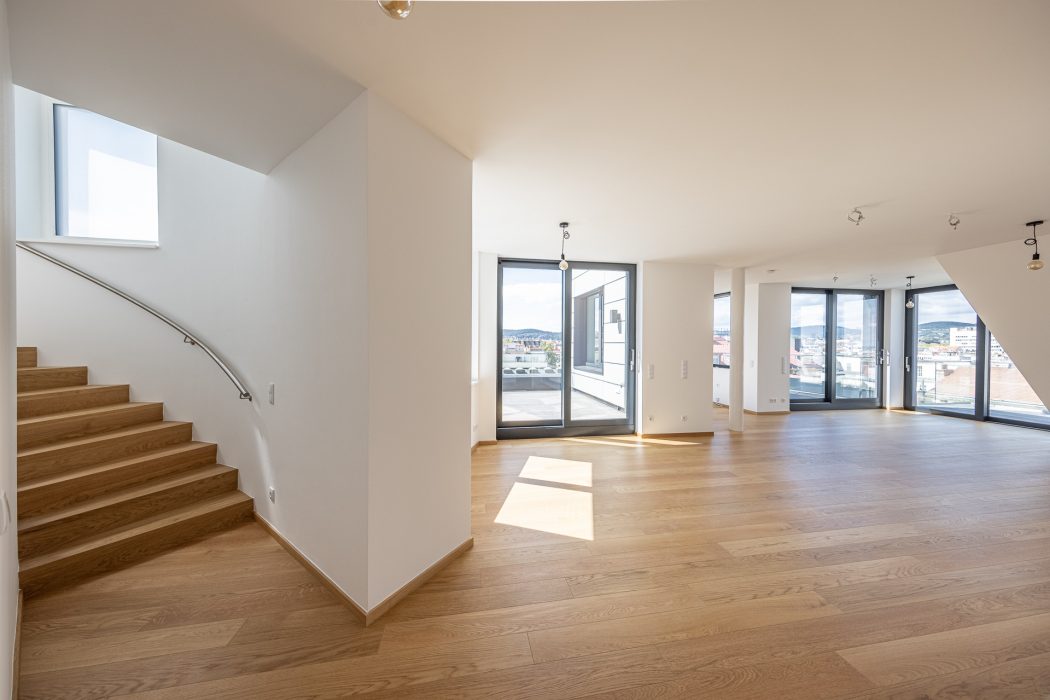 Innenraumansicht eines modernen, leeren Wohnraums mit Holzfußboden, großen Fenstern und einer Treppe mit Metallgeländer links.