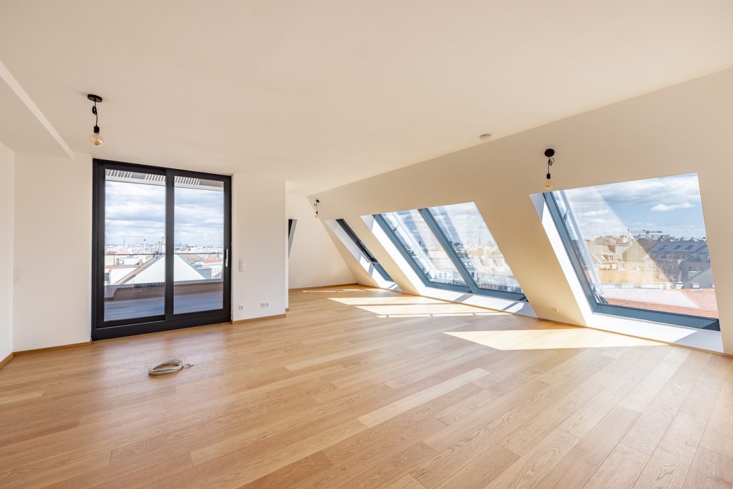 Helles, leeres Zimmer mit Holzboden und schrägen Dachfenstern, die Ausblick auf eine städtische Dachlandschaft bieten.