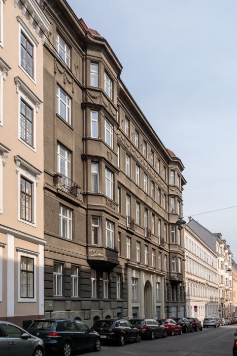 Straßenansicht eines mehrstöckigen, historischen Gebäudes mit Erkern und dekorativen Elementen, davor parken Autos.