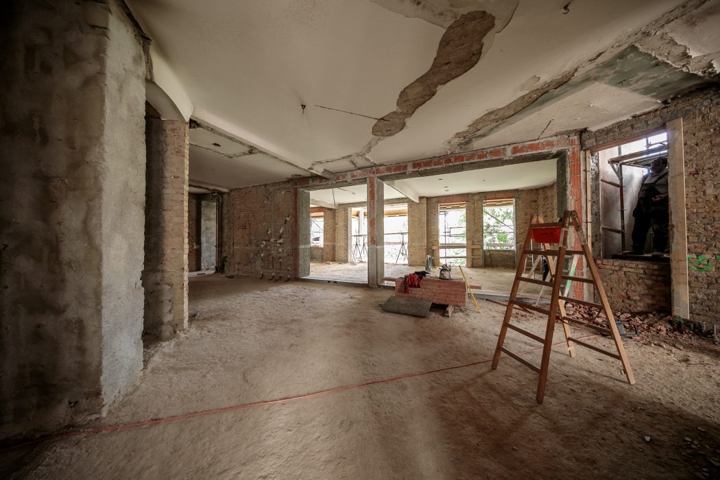 Ein unvollständiger Raum im Bau mit unverputzten Ziegelwänden, einem Leiter in der Mitte und zwei Bauarbeitern bei einem Fenster rechts.