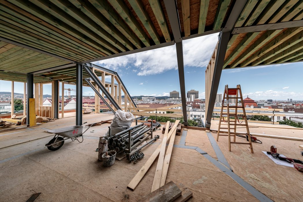 Baustelle mit Holzbalken, Metallstützen, Leiter, Schubkarre und Werkzeugen, Blick auf Stadtlandschaft im Hintergrund.
