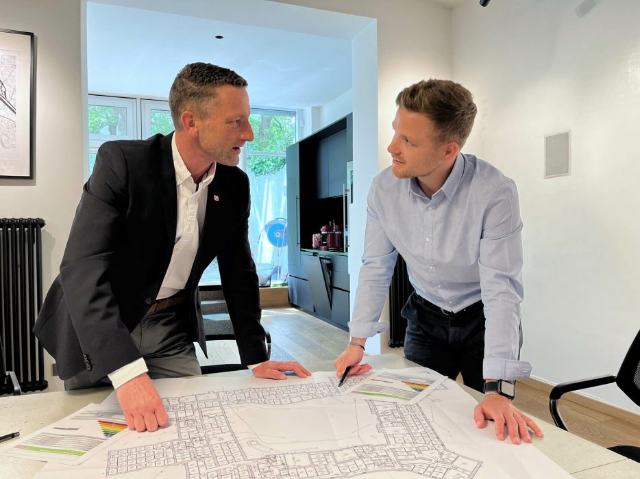 Zwei Männer stehen an einem Tisch in einem modernen Büro und diskutieren über einen Stadtplan, der auf dem Tisch liegt.