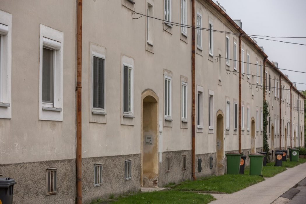 Reihenhausfassade mit mehreren weißen Fenstern und Türen, Mülltonnen entlang des Gehwegs und Stromleitungen über der Straße.