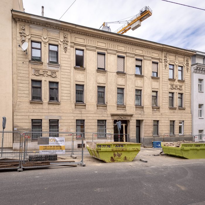 Ein älteres vierstöckiges Gebäude mit Sandsteinfassade, Bauarbeiten im Vordergrund, Metallzäune und grüne Abfallcontainer auf der Straße.