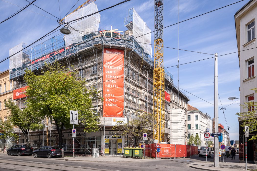 Straßenkreuzung mit Baustelle. Gebäude mit Baugerüst, großer Kran, orangenes Schild auf Deutsch, Autos und Verkehrsschilder sichtbar.