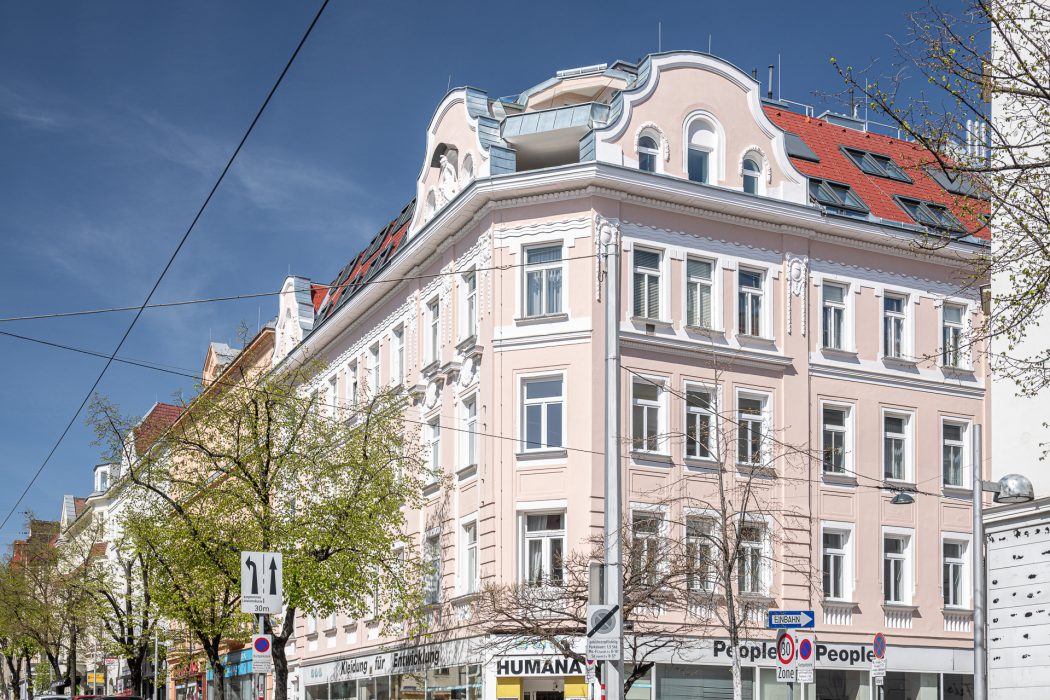 Ein rosa-weißes historisches Gebäude an einer Straßenecke mit mehreren Etagen und Geschäften im Erdgeschoss unter klarem, blauem Himmel.