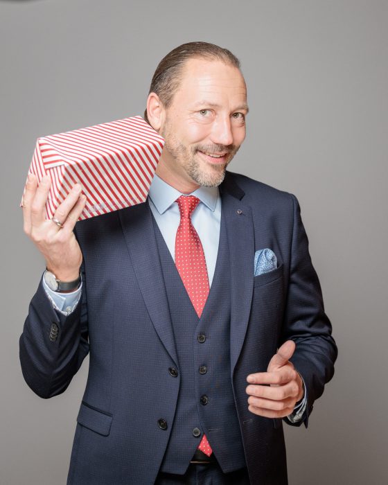 Ein Mann in einem dunkelblauen Anzug mit roter Krawatte hält ein rot-weiß gestreiftes Geschenk und lächelt in die Kamera.
