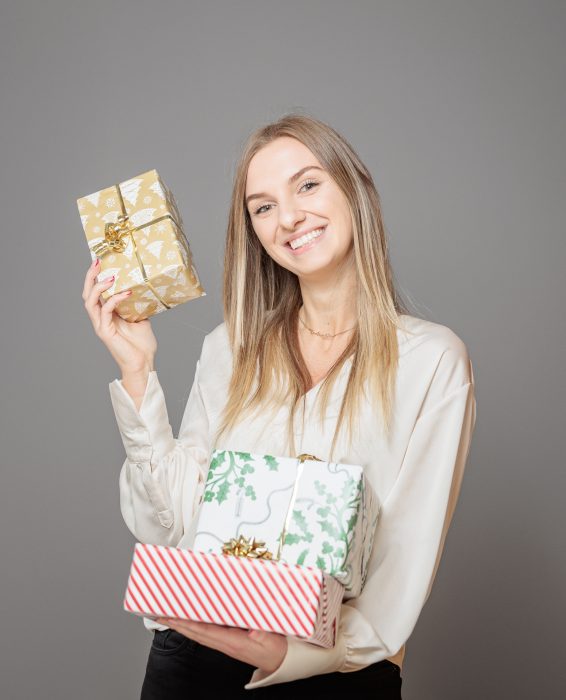 Eine lächelnde Frau in weißer Bluse hält drei Geschenke mit verschiedenen Geschenkpapiermustern und goldenen Schleifen in den Händen.