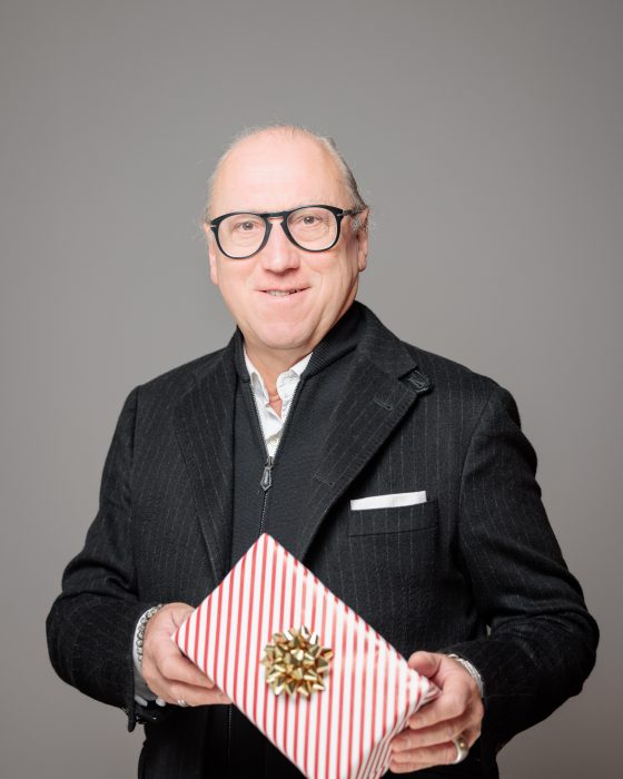 Ein älterer Mann mit Brille hält ein rot-weiß gestreiftes Geschenk mit einer goldenen Schleife, trägt einen schwarzen Anzug.