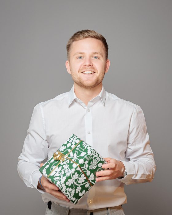 Ein lächelnder Mann in einem weißen Hemd hält ein grün-weiß gemustertes Geschenk mit goldener Schleife vor einem grauen Hintergrund.