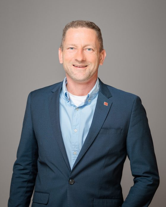 Ein lächelnder Mann in einem blauen Anzug und hellblauem Hemd steht vor einem grauen Hintergrund.