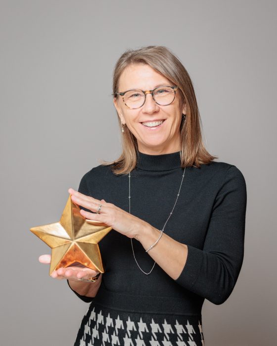 Eine Frau mit Brille und schulterlangem Haar hält lächelnd einen großen goldenen Stern in den Händen.