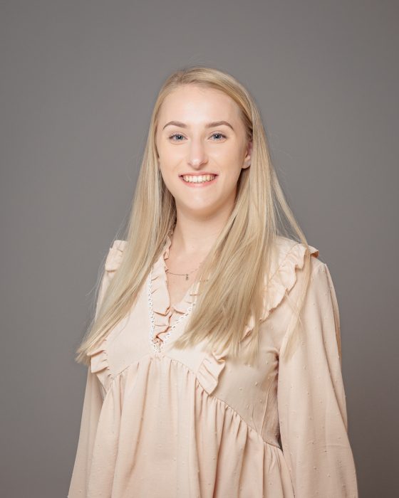 Eine lächelnde Frau mit langen blonden Haaren trägt eine beigefarbene Bluse mit Rüschen und steht vor einem neutralen grauen Hintergrund.