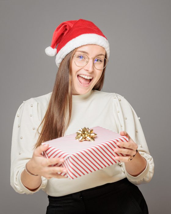 Eine lachende Frau mit Brille trägt eine rote Weihnachtsmütze und hält ein weiß-rot gestreiftes Geschenk mit einer goldenen Schleife.