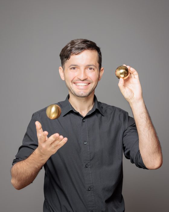 Ein lächelnder Mann in einem schwarzen Hemd jongliert mit goldenen Kugeln vor einem grauen Hintergrund.