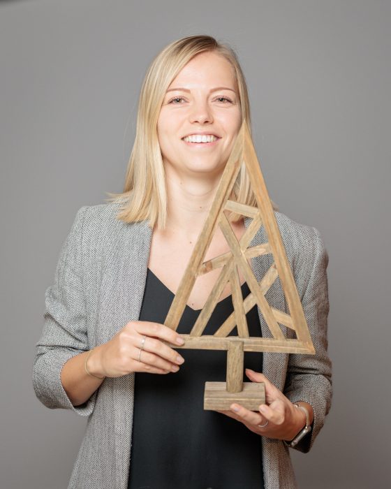Eine lächelnde Person mit blondem Haar hält eine hölzerne, geometrische Baumskulptur vor einem grauen Hintergrund.