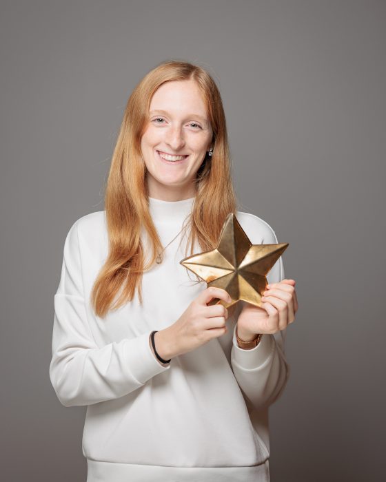 Person mit langen, roten Haaren hält einen goldenen Stern und lächelt. Sie trägt ein weißes Sweatshirt vor grauem Hintergrund.