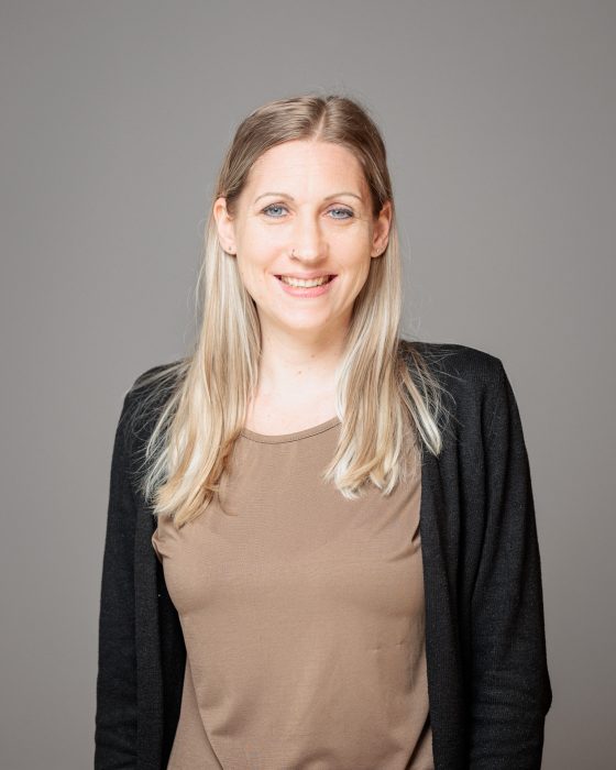 Eine lächelnde Frau mit langen, blonden Haaren trägt ein braunes Oberteil und eine schwarze Strickjacke vor einem grauen Hintergrund.