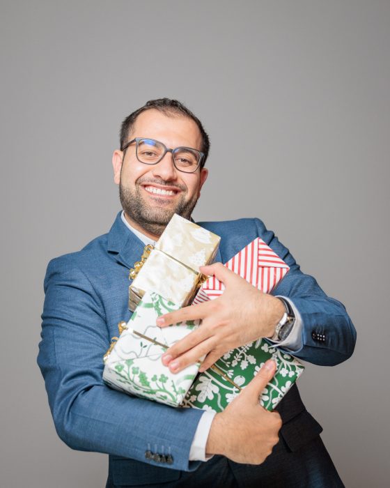 Ein lächelnder Mann im Anzug hält mehrere dekorativ verpackte Geschenke in den Armen vor einem neutralen Hintergrund.