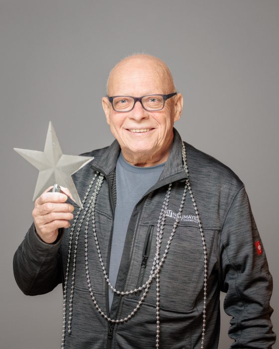 Ein älterer Mann mit Brille hält einen Stern und trägt silberne Perlenketten. Er lächelt und trägt eine dunkelgraue Jacke.