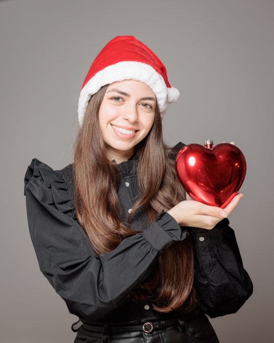 Eine Frau mit langem Haar trägt eine Weihnachtsmütze und hält ein rotes, herzförmiges Ornament, lächelnd vor neutralem Hintergrund.