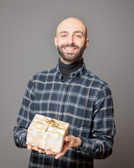 Ein lächelnder Mann in kariertem Hemd hält ein festlich verpacktes Geschenk mit goldenen Mustern und Schleife vor einem grauen Hintergrund.