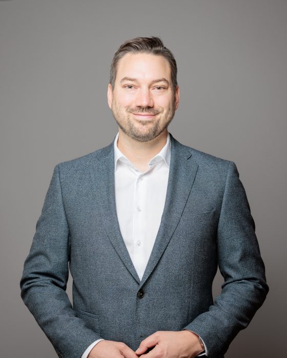 Ein Mann in Anzug und Hemd steht vor einem grauen Hintergrund und lächelt in die Kamera.